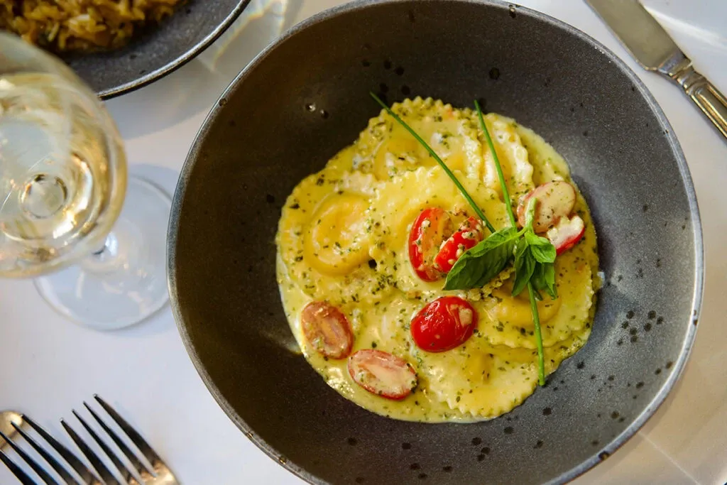 A delectable dish of ravioli is elegantly presented in a dark bowl, topped with fresh cherry tomatoes, a sprig of chives, and a basil garnish, all lightly drizzled with a creamy herb sauce. The setting is part of Le Crystal, a renowned wedding venue in the category of Wedding Venues on Mariage Elegant, Canada. In the background, a glass of white wine and polished silverware contribute to the sophisticated dining ambiance typical of wedding celebrations.
