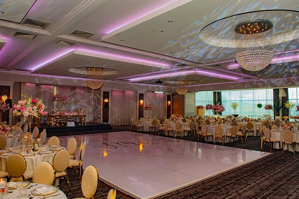 The image showcases an elegant wedding venue by Le Crystal, categorized under wedding venues on Mariage Elegant, Canada. The spacious ballroom is beautifully decorated, featuring a large, polished dance floor in the center. Surrounding the dance floor are round tables elegantly set with fine china, gold-rimmed glasses, and cream-colored chairs. Soft, ambient lighting casts a warm glow throughout the room, while opulent chandeliers hang from the ceiling, enhancing the luxurious atmosphere. Floral arrangements with pink and white flowers adorn the tables and venue, adding a touch of freshness and romance to the sophisticated decor.
