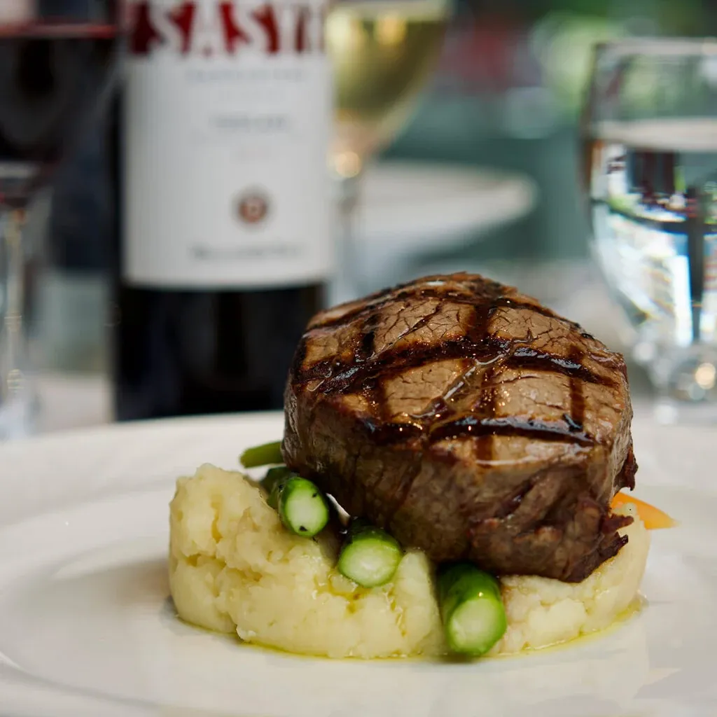 A gourmet dish is elegantly plated at Le Crystal, a premier wedding venue in Canada. The image showcases a perfectly grilled filet mignon resting on a bed of creamy mashed potatoes, garnished with tender asparagus spears. In the background, there is a bottle of wine and a filled wine glass, highlighting the luxurious dining options available at this venue. A clear glass of water is also present, completing the sophisticated table setting. This presentation reflects the fine dining experience that couples can expect when choosing Le Crystal for their wedding celebrations.