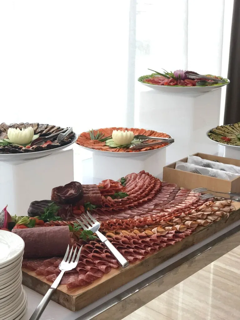 The image showcases an elegant charcuterie spread at Le Crystal, a prominent wedding venue listed under the wedding venues category for Mariage Elegant in Canada. The table is artfully arranged with an array of sliced meats including salami and prosciutto, accented with garnishes such as herbs and vegetables. Elevating the presentation are plates on pedestals in the background, featuring additional charcuterie selections and decorative vegetables. White plates and serving forks are neatly placed in the foreground, ready for guests to use, enhancing the venue's sophisticated and inviting atmosphere.