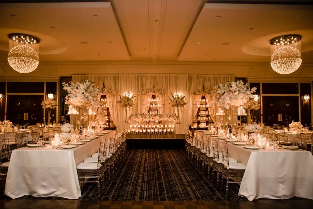 The image showcases an elegant wedding venue by Le Crystal, categorized under Wedding Venues, featured on Mariage Elegant in Canada. The luxurious setting displays a beautifully decorated banquet hall with a sophisticated ambiance. Long rectangular tables are adorned with crisp white tablecloths, gold tableware, and soft candlelight, creating a warm and inviting glow. Stunning floral arrangements of white and blush flowers elegantly tower above the tables. In the background, a grand floral centerpiece is prominently displayed on a raised platform, surrounded by shimmering candles, enhancing the romantic atmosphere. The ceiling features exquisite, spherical chandeliers that further elevate the venue's grandeur, making it an ideal setting for an upscale wedding celebration.
