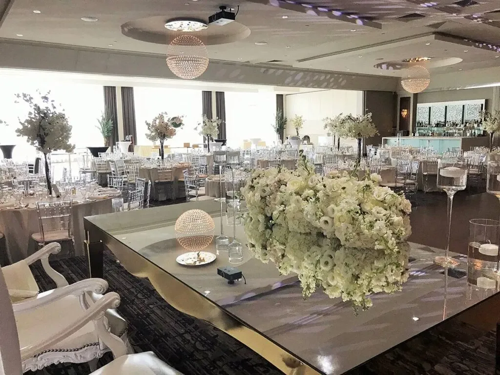This image showcases a sophisticated wedding venue by Le Crystal, categorized under Wedding Venues on Mariage Elegant, Canada. The spacious room is elegantly decorated, featuring an abundance of round tables set for a wedding reception. The tables are adorned with luxurious white tablecloths and topped with ornate floral centerpieces, primarily consisting of white flowers. The venue's ambient lighting includes spherical chandeliers that cast a warm glow over the room. In the foreground, a large mirrored table is lavishly decorated with another opulent floral arrangement, adding a touch of elegance. The room also features chiavari chairs, enhancing the upscale atmosphere, while a modern bar is visible in the background, providing a chic touch to the setting.