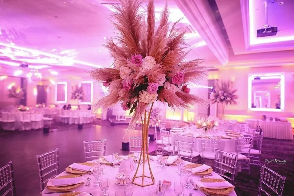 The image showcases a beautifully decorated wedding venue by Le Crystal, a featured vendor in the Wedding Venues category on Mariage Elegant, Canada. The room is elegantly lit with a soft pink and purple glow, adding a romantic ambiance. At the center is a round table adorned with a tall, golden stand centerpiece overflowing with pampas grass, pink roses, and white flowers. The tables are set with white tablecloths, neatly arranged glassware, and plates with golden napkins, complemented by transparent chiavari chairs. The background features more tables similarly arranged, highlighting the venue's spaciousness and luxurious design.