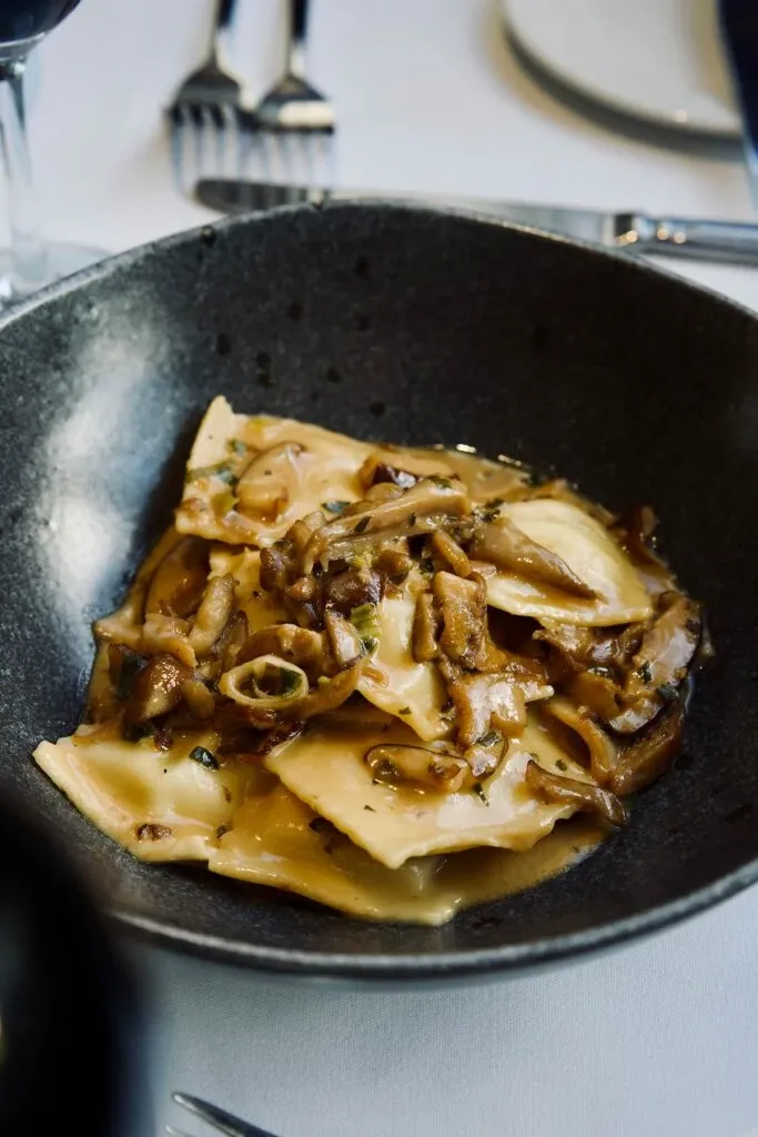 A close-up of a gourmet pasta dish is presented in a dark bowl, featuring ravioli topped with a savory mushroom sauce. The elegant dining setup is part of Le Crystal, highlighting their exquisite catering as a premier wedding venue in Canada. The table is set with shiny silver cutlery and a pristine white tablecloth, showcasing the attention to detail and luxurious dining experience provided by this vendor.