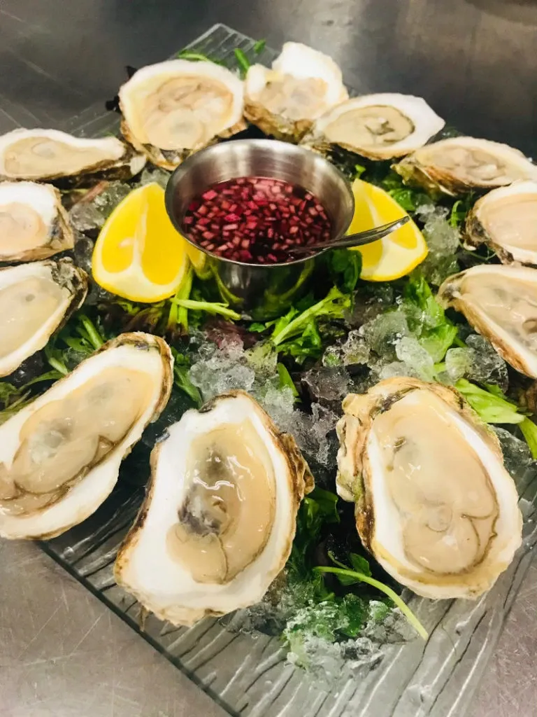The image showcases a beautifully arranged seafood platter featuring fresh oysters served on a bed of crushed ice. The oysters are opened and neatly placed in a circle around a central metal sauce container filled with a vibrant red mignonette sauce. Two slices of lemon are strategically placed on the platter for added freshness. Greenery is used as an additional garnish, enhancing the presentation. This dish, likely part of Le Challenger's catering offerings, exemplifies the elegance suitable for weddings at this Canadian wedding venue.
