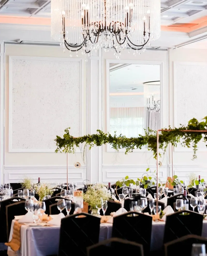 The image showcases an elegantly decorated wedding venue at Le Challenger, categorized under Wedding Venues on Mariage Elegant, Canada. The room features stunning crystal chandeliers hanging from a coffered ceiling, casting a soft, romantic light. Below, tables are beautifully set with white linens and black chairs, each adorned with delicate glassware and intricate floral centerpieces featuring lush greenery. The overall ambiance is sophisticated and inviting, perfect for a memorable wedding celebration.