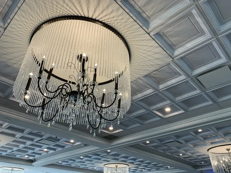 The image features an elegant wedding venue, Le Challenger, which falls under the category of Wedding Venues. The ceiling is adorned with luxurious chandeliers draped in cascading crystal strands, contributing to a sophisticated atmosphere. The chandeliers' lights softly illuminate the coffered ceiling, enhancing the venue’s refined architectural details. This venue is ideal for hosting an elegant wedding reception, offering a stunning visual ambiance for special occasions.