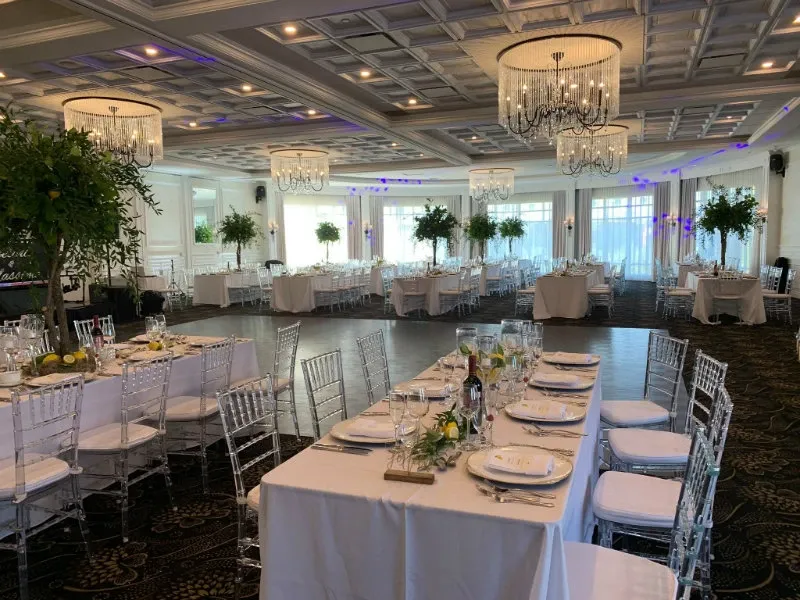 The image showcases a beautifully arranged wedding venue at Le Challenger, categorized under Wedding Venues. The spacious room features elegantly set tables adorned with crisp white linens and floral centerpieces, creating an inviting atmosphere. Clear, modern chairs complement the sophisticated décor, while large, ornate chandeliers hang from the intricately designed ceiling, casting a warm glow over the scene. The room is bathed in natural light streaming through expansive windows, highlighting the tasteful ambiance, ideal for weddings and receptions.