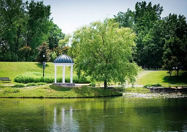 The image showcases Le Challenger, a picturesque wedding venue surrounded by lush greenery in Canada. Central to the scene is a charming white gazebo with a domed roof, perfect for a romantic ceremony. It is situated by a serene pond, reflecting the vibrant trees and blue sky. The peaceful setting is complemented by manicured lawns, inviting benches, and distant trees, offering an idyllic backdrop for memorable wedding photos.