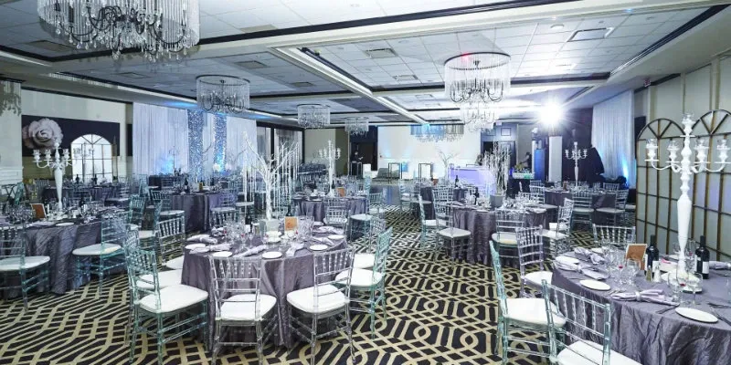 The elegantly decorated interior of Le Challenger, a renowned wedding venue in Canada, features a spacious banquet hall adorned with crystal chandeliers and tastefully set round tables. Each table is covered with luxurious gray tablecloths and neatly arranged silverware, with clear Chiavari chairs enhancing the sophisticated ambiance. The room is softly lit, creating a romantic atmosphere ideal for weddings, while tall birch-like centerpieces and delicate lighting add a touch of elegance to the setting. The attention to detail at Le Challenger makes it a sought-after choice for couples planning their special day.
