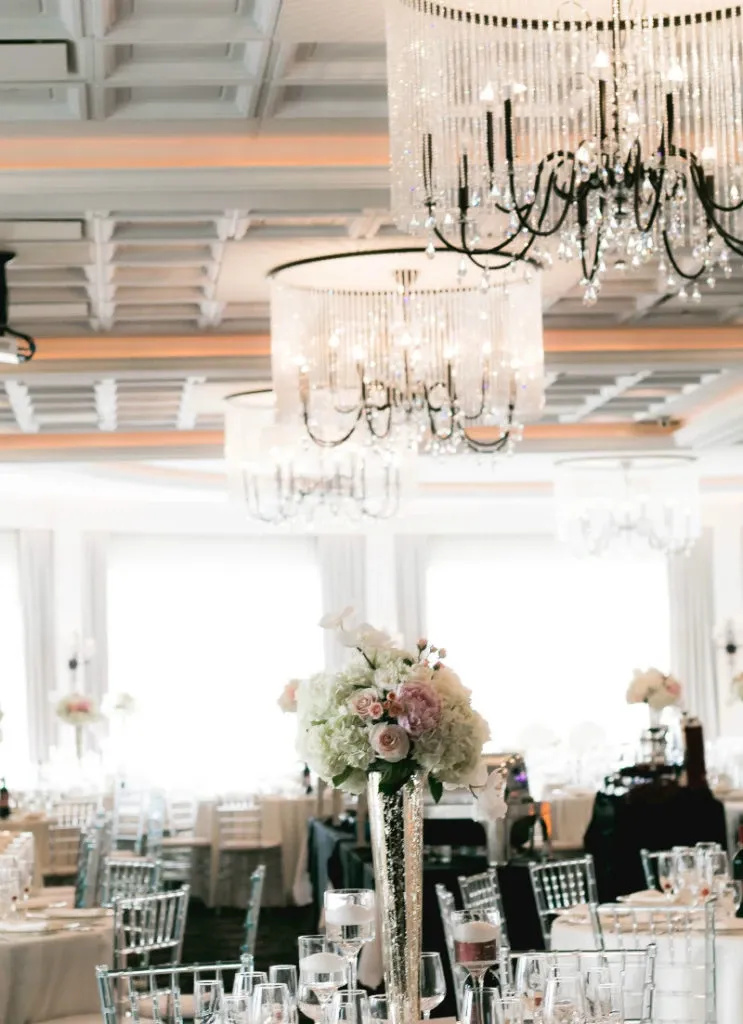 The image showcases an elegant wedding venue by Le Challenger, categorized under Wedding Venues, featured on Mariage Elegant, Canada. The venue is beautifully decorated with an opulent interior featuring multiple crystal chandeliers that gracefully illuminate the room. Guests are seated at tables adorned with exquisite floral centerpieces in soft pink and white hues. The room, filled with natural light streaming through large windows, creates a romantic ambiance perfect for weddings. Transparent Chiavari chairs and elegantly set tables add a sophisticated touch to the celebration.