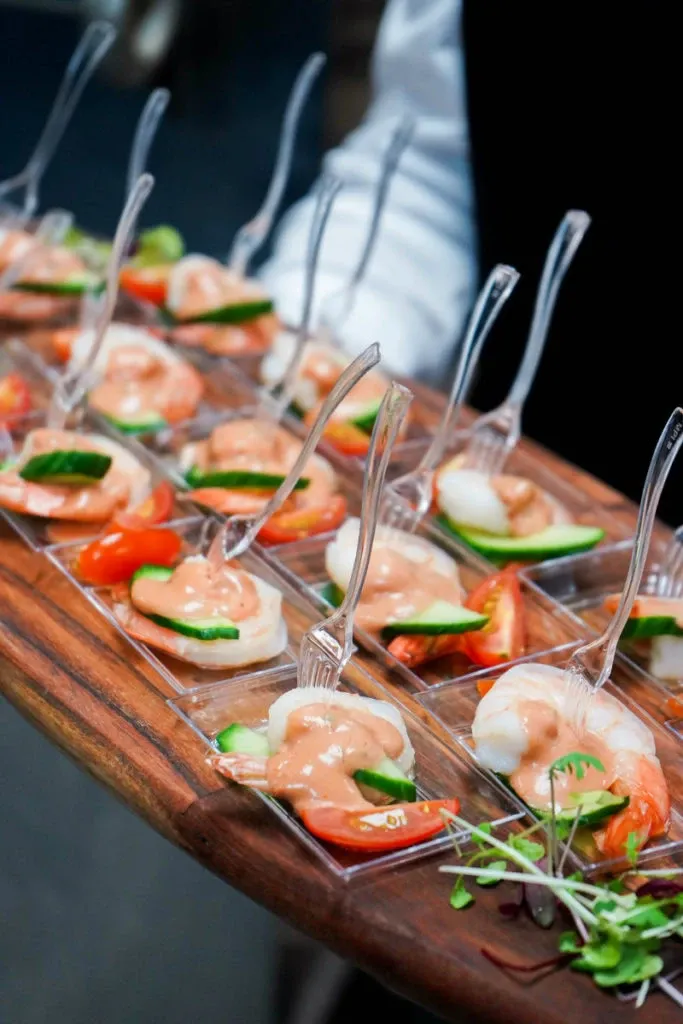 In this image from Le Challenger, a premier wedding venue in Canada listed on Mariage Elegant, a beautifully arranged tray of appetizers is showcased. Each portion features fresh shrimp paired with a dollop of creamy sauce, garnished with a sliver of cucumber and a cherry tomato slice, all elegantly presented in clear, individual serving spoons. The appetizers are artistically arranged on a wooden serving board, highlighting the venue’s commitment to exquisite catering and attention to detail for wedding events.