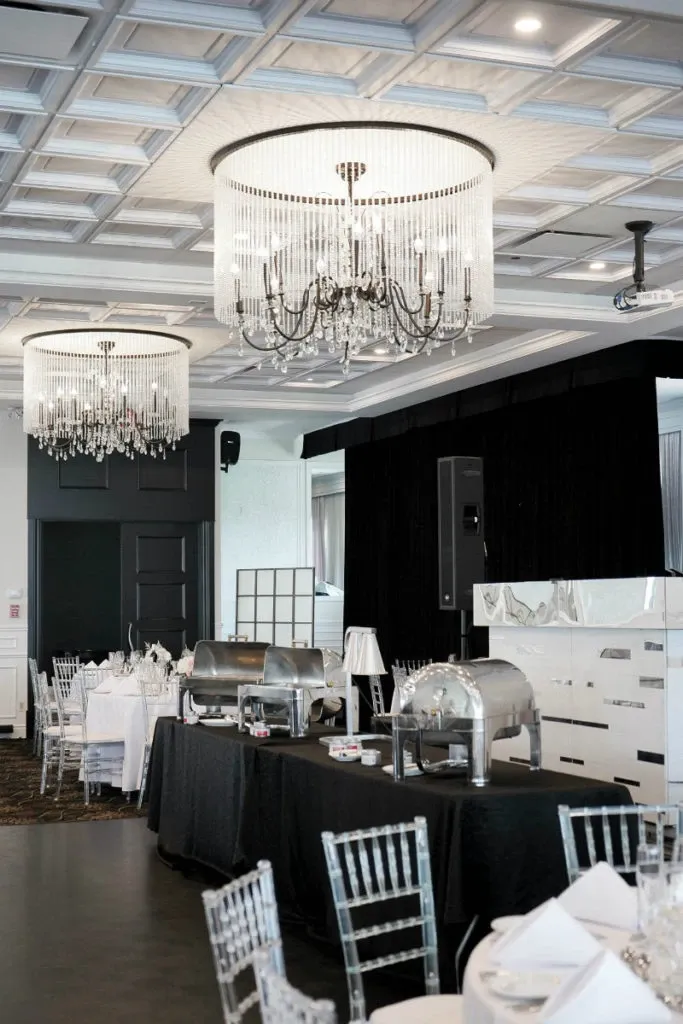 This image features an elegant wedding venue, Le Challenger, categorized under Wedding Venues on the Mariage Elegant site in Canada. The venue showcases a sophisticated banquet hall adorned with two large, ornate chandeliers hanging from a detailed coffered ceiling. Below are neatly arranged round dining tables covered with pristine white tablecloths and set with elegant dinnerware, surrounded by clear Chiavari chairs. A buffet station with covered chafing dishes is prominently displayed, adding to the formal atmosphere. The venue exudes a luxurious and stylish setting, ideal for a memorable wedding celebration.