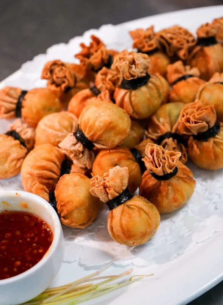 The image shows a beautifully arranged platter of golden-brown, intricately wrapped appetizers on a white plate, accompanied by a small bowl of dipping sauce with a rich red hue, hinting at a spicy flavor. The appetizers are artfully tied with dark, delicate strips, resembling small pouches, offering a sophisticated touch to wedding catering. This presentation is part of Le Challenger's offerings, highlighting their expertise as a premier wedding venue under the category of Wedding Venues in the Mariage Elegant vendor listing in Canada.