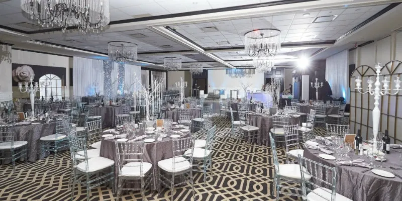 The image shows the elegant interior of Le Challenger, a premier wedding venue in Canada, set up for a special event. The spacious room features numerous round tables draped in luxurious gray tablecloths, each adorned with pristine white plates and gleaming silverware, suggesting a formal dining setup. Stylish transparent chairs surround the tables, enhancing the modern aesthetic. Overhead, crystal chandeliers hang from the ceiling, casting a soft, inviting light over the venue. The floor displays a striking geometric pattern that complements the sophisticated decor. In the background, white drapes and decorative lighting complete the romantic and airy ambiance, making Le Challenger an ideal choice for weddings.