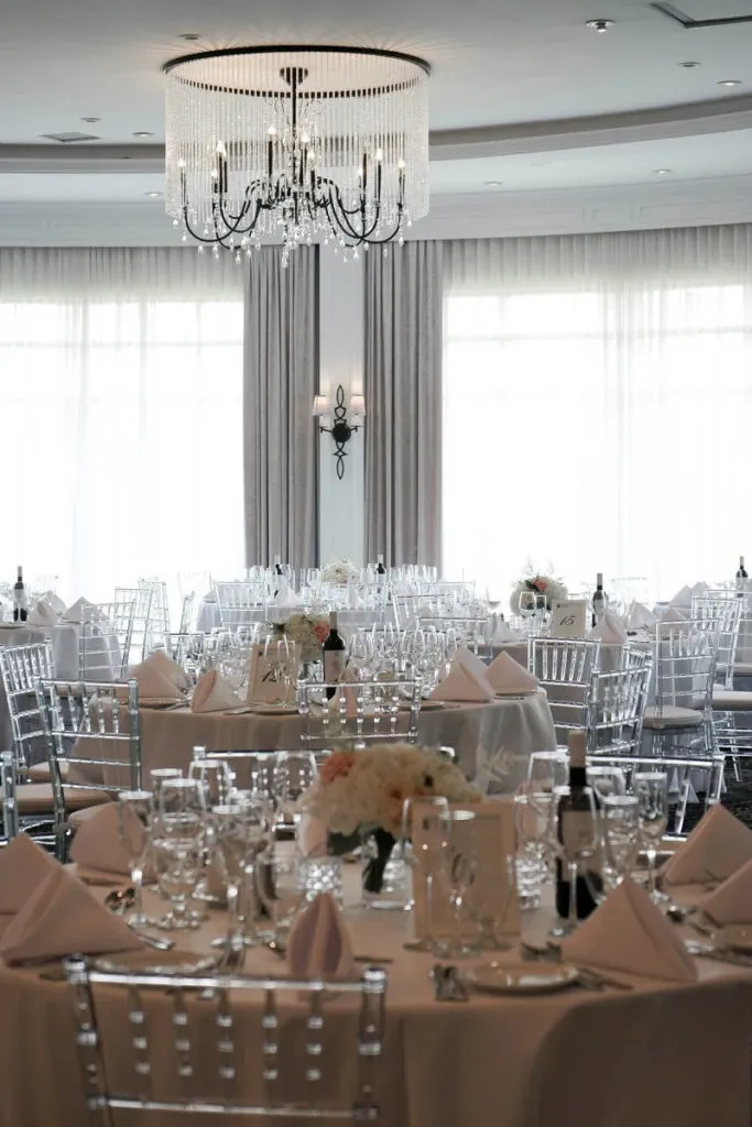 The image showcases the elegant interior of Le Challenger, a premier wedding venue featured on Mariage Elegant, Canada. The spacious room is beautifully set for a wedding reception, with round tables draped in white linens. Each table is elegantly arranged with wine glasses, napkin settings, and delicate floral centerpieces. The venue exudes sophistication with its crystal chandelier hanging from the ceiling, complementing the soft natural light streaming in from large windows dressed with long curtains. Chiavari chairs add a modern touch, enhancing the venue's luxurious ambiance.