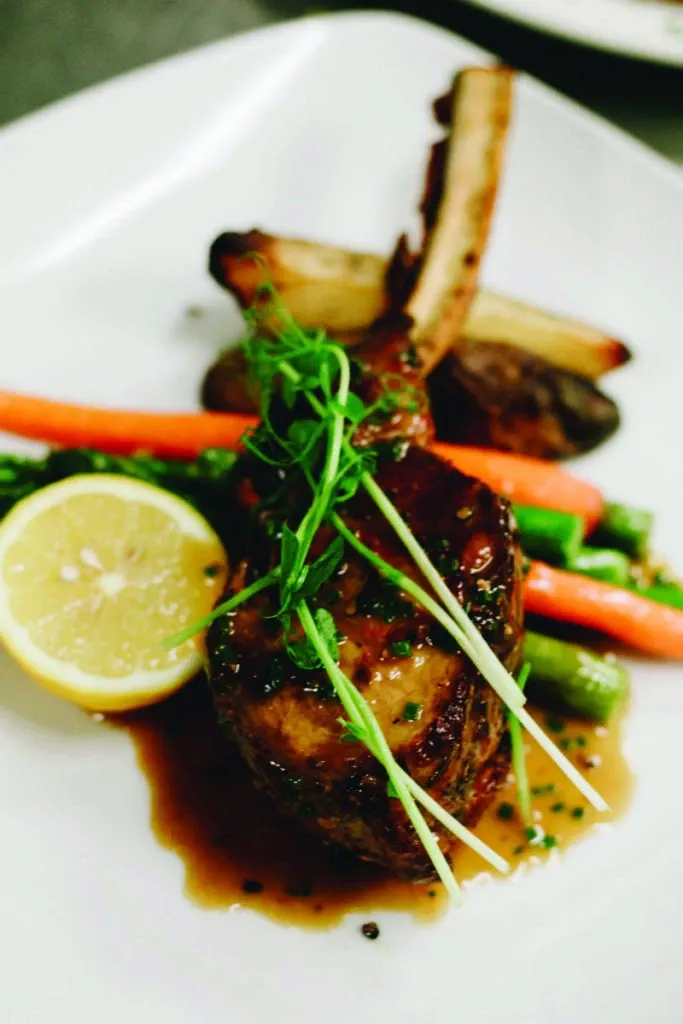 A beautifully presented gourmet dish from Le Challenger, a top-tier wedding venue in the Mariage Elegant network, Canada. The plate features a perfectly seared lamb chop drizzled with a rich, savory glaze, garnished with fresh greens. Accompanying the lamb are vibrant sides including roasted carrots, asparagus, and a lemon wedge, artfully arranged to enhance the dining experience. This elegant culinary presentation highlights the exquisite catering services offered by this wedding venue, ensuring a memorable experience for guests.