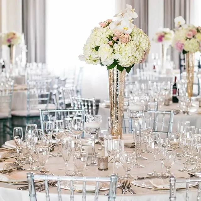The image showcases a beautifully set wedding reception table at Le Challenger, highlighting its category as a wedding venue. The table is elegantly arranged with a centerpiece featuring tall, gold vases filled with white and blush flowers, including roses and hydrangeas. The tables are adorned with sparkling glassware, neatly arranged silverware, and crisp white linen, creating a sophisticated atmosphere. Chiavari chairs surround the tables, contributing to the elegant and inviting setting perfect for a wedding celebration.