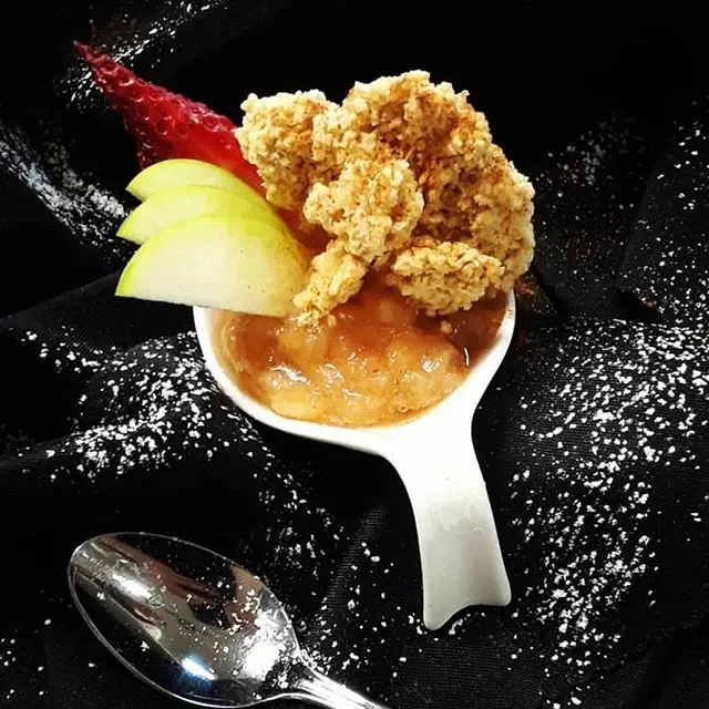 A beautifully presented dessert at Le Challenger, a premium wedding venue in Canada, is elegantly displayed on a black cloth backdrop. The dish features a creamy compote topped with a generous oat crumble. Artful garnishes include thin apple slices and a perfectly ripe strawberry, adding color and freshness. A dusting of powdered sugar surrounds the dessert, enhancing its visual appeal. A polished silver spoon lies in the foreground, suggesting readiness for tasting. This representation showcases the venue's attention to detail and exquisite catering options, ideal for wedding celebrations hosted at Le Challenger.