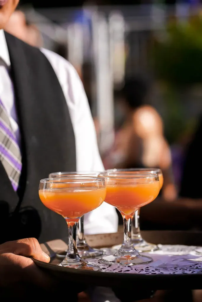 A server dressed in formal attire holds a tray with three elegant cocktail glasses containing a peach-colored beverage, likely a signature drink for a wedding reception. The setting appears to be a lively outdoor event, with blurred figures and lush greenery visible in the background, creating a festive atmosphere. This image represents the exquisite service offered by Le Challenger, a wedding venue listed in the Mariage Elegant vendor directory under the Wedding Venues category in Canada.