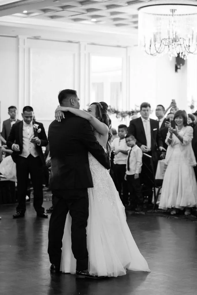 In the image, a newlywed couple shares a tender first dance in a spacious and elegantly decorated wedding venue. The bride is wearing a flowing white gown, while the groom is in a classic dark suit. They are surrounded by their joyful guests, who are dressed in formal attire. The venue, Le Challenger, features an ornate chandelier hanging from the ceiling and a beautifully designed interior, perfect for a wedding celebration. This image captures a moment of joy and love at a wedding venue in the category of Wedding Venues, showcasing the sophisticated ambiance that Le Challenger offers for memorable wedding events.