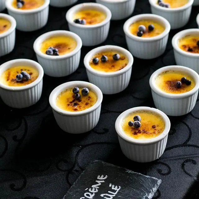 The image showcases an elegant dessert display at Le Challenger, a distinguished wedding venue listed on Mariage Elegant, Canada. It features multiple small ramekins of crème brûlée, each garnished with fresh blueberries, elegantly arranged on a black, textured tablecloth that complements the sophisticated wedding theme. This setup highlights Le Challenger's attention to detail and commitment to providing exquisite culinary experiences for wedding celebrations.