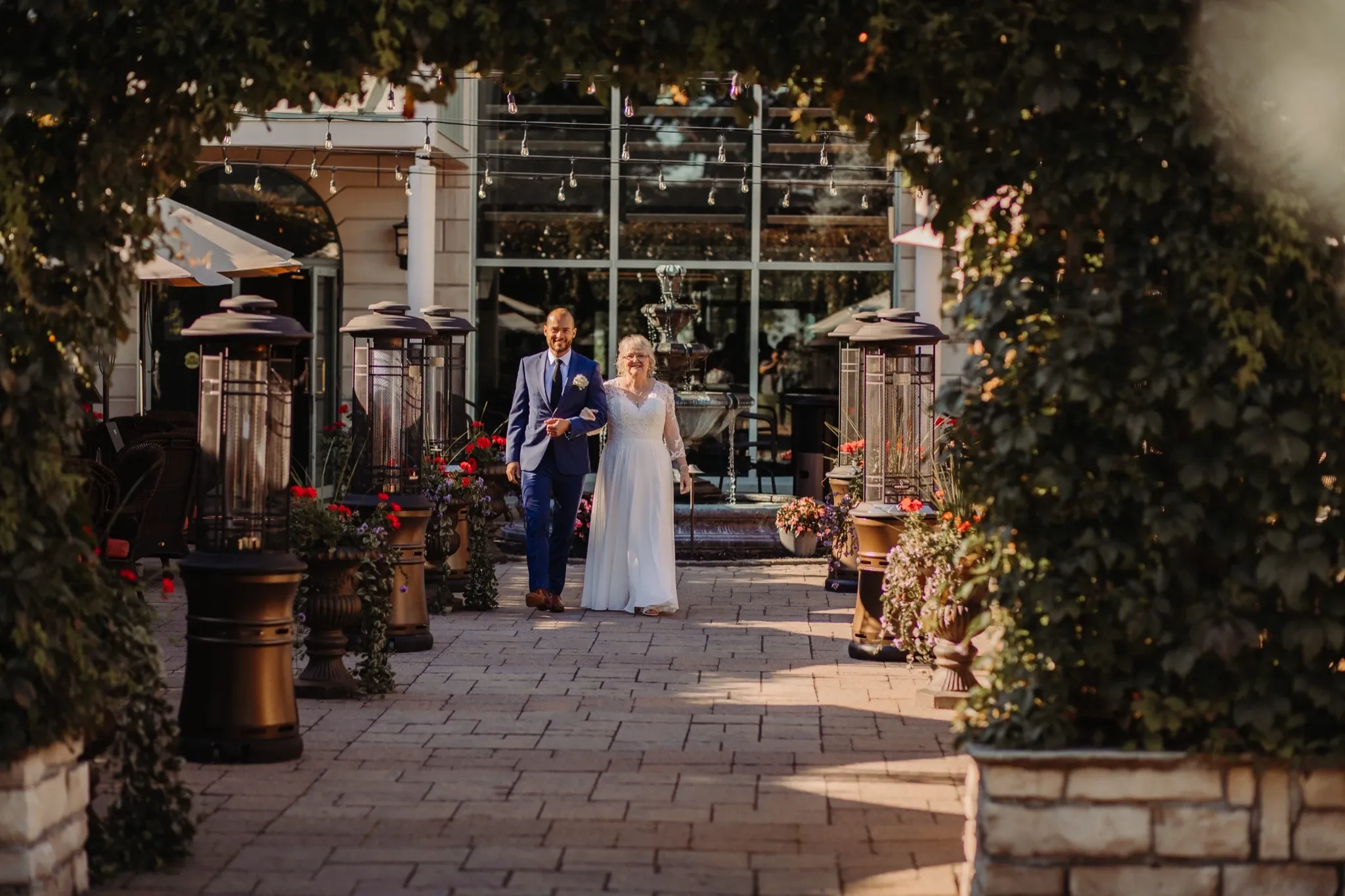 A bride and groom walk together down a beautifully landscaped garden pathway at Auberge Godefroy, a wedding venue. The paved path is lined with lush greenery and vibrant flowers in elegant pots, leading towards a building with large glass windows. Outdoor heaters and decorative lighting enhance the romantic atmosphere, creating a perfect setting for a wedding ceremony or reception. The couple looks serene and happy, embodying the elegance and charm of a memorable wedding day at this Canadian venue.