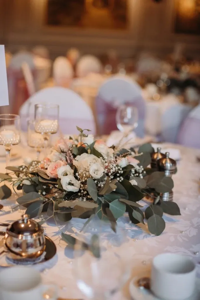 The image showcases a beautifully arranged wedding reception table at Auberge Godefroy, a wedding venue in Canada. The table is elegantly set with a centerpiece featuring an assortment of fresh flowers, including white roses and delicate pink blossoms, accented by lush green leaves and baby's breath. Each place setting includes polished silverware and glassware, adding a touch of sophistication. Soft, ambient lighting complements the decor, creating an inviting and romantic atmosphere typical for weddings hosted at this venue.