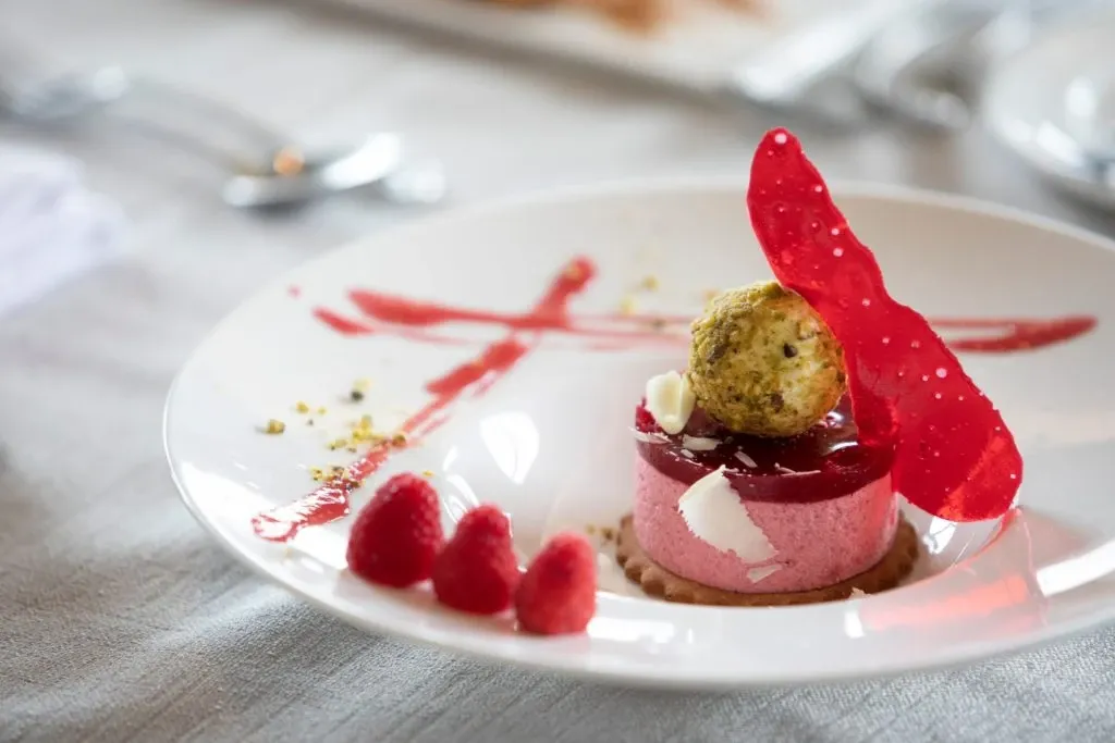 The image showcases a beautifully plated dessert, suitable for a wedding reception at Auberge Godefroy, a premier wedding venue in Canada. The centerpiece is an elegantly arranged raspberry mousse cake topped with a delicate pistachio tuile and garnished with a vibrant red sugar decoration. Fresh raspberries and a drizzle of raspberry sauce are artfully placed on the white plate, creating a visually appealing contrast. The dessert is set on a finely textured white tablecloth, with cutlery subtly in the background, enhancing the sophisticated and elegant ambiance typical of wedding venues like Auberge Godefroy.