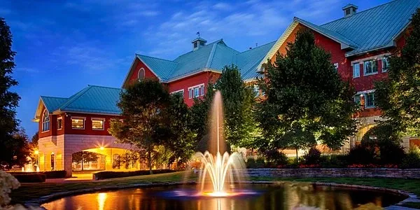 The image showcases Auberge Godefroy, a premier wedding venue in Canada, part of the Mariage Elegant listing. The venue features an impressive, large building with a distinctive red facade and green, gabled roofs, set against a stunning twilight sky. In the foreground, there's a beautifully lit fountain surrounded by a manicured garden, adding a touch of elegance and romance to the setting. This venue offers a picturesque backdrop ideal for wedding ceremonies and receptions.
