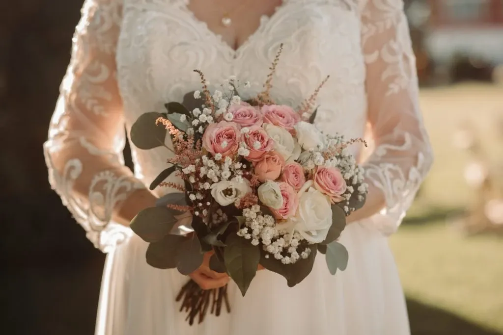 A bride holding a beautifully arranged bouquet of pink and white roses with accents of baby's breath and eucalyptus, set against the background of a sunlit outdoor setting. Her dress features intricate lace details on the sleeves, suggesting an elegant and sophisticated wedding theme. This image represents Auberge Godefroy, a distinguished wedding venue in Canada, ideal for couples seeking a charming and picturesque location for their special day.
