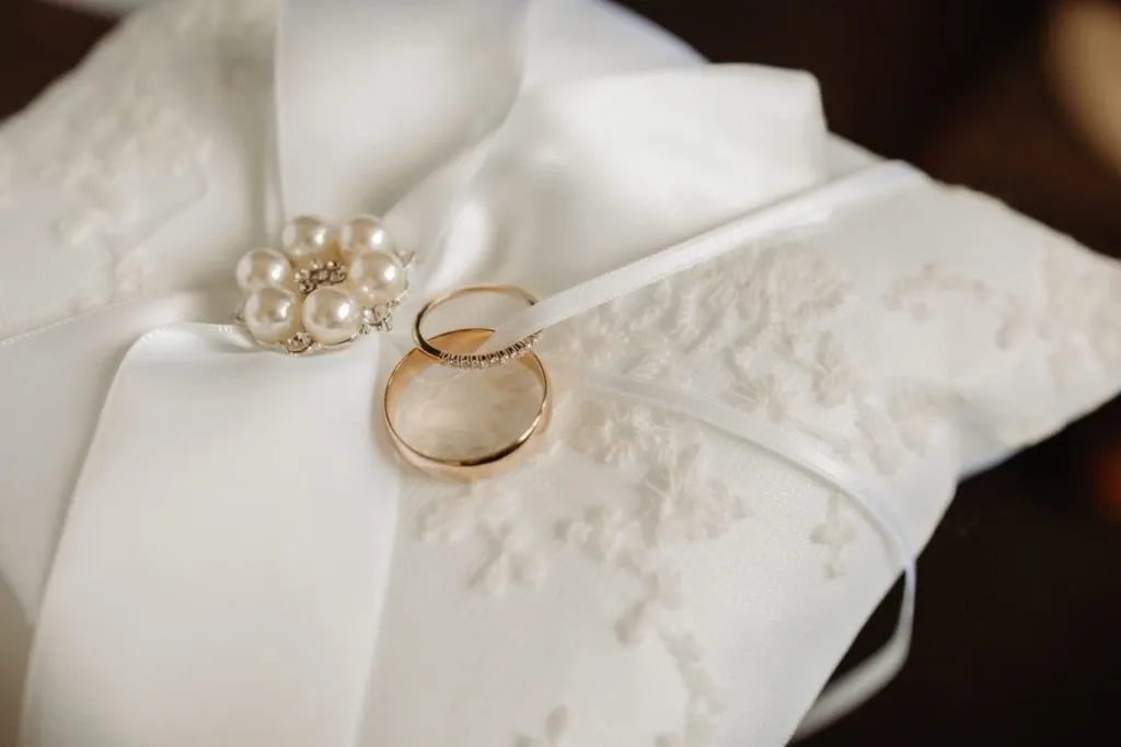 The image features two elegant wedding rings resting on a delicate, white satin pillow adorned with intricate lace patterns. A decorative brooch with pearls and rhinestones enhances the pillow’s sophistication, tied with a soft satin ribbon. The image is associated with Auberge Godefroy, highlighting its offerings in the Wedding Venues category on Mariage Elegant, Canada. This visually symbolizes the elegance and attention to detail expected from a premier wedding venue.