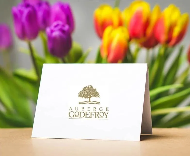 The image features a folded card on a wooden surface with the gold-embossed logo of Auberge Godefroy, a renowned wedding venue in Canada. In the background, vibrant tulips in shades of purple, pink, and yellow provide a colorful and elegant touch, highlighting the venue's association with beauty and sophistication. This setting underscores Auberge Godefroy's appeal as an ideal location for weddings, offering a blend of elegance and charm.