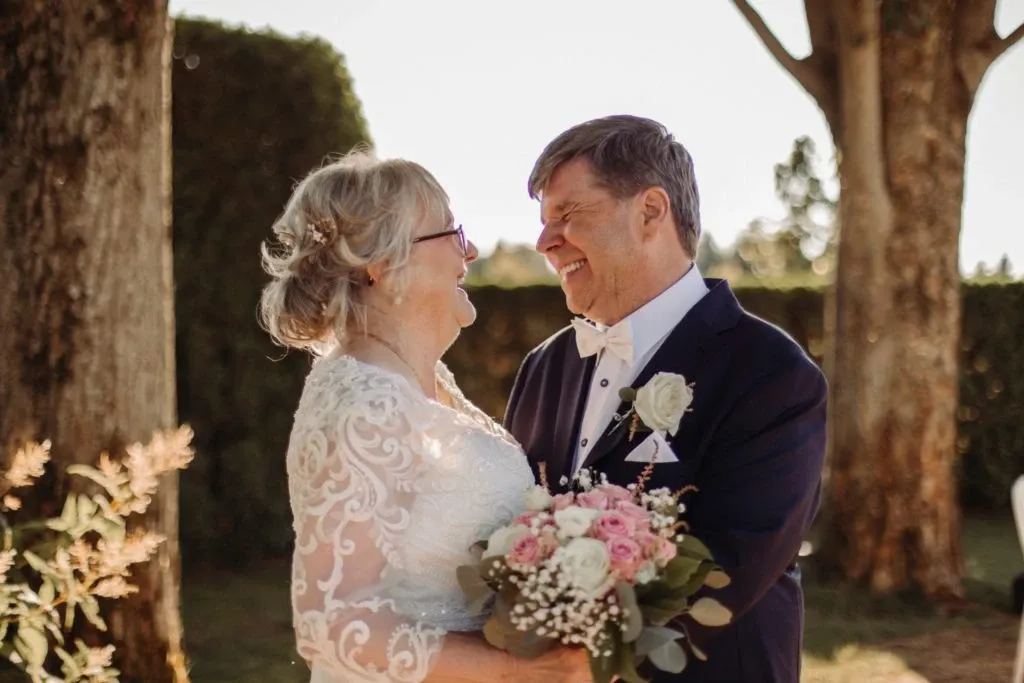 A happy couple smiles at each other on their wedding day at Auberge Godefroy, nestled in a picturesque outdoor setting. The bride, wearing a beautifully detailed lace gown, holds a bouquet of pink and white flowers accented with greenery and baby's breath. The groom is dressed in a classic dark suit with a white shirt and bow tie, adorned with a white rose boutonniere. Tall trees and manicured hedges provide a serene and elegant backdrop, epitomizing the charm of this wedding venue. Ideal for couples seeking a romantic and intimate atmosphere, Auberge Godefroy offers a perfect setting for memorable wedding celebrations in Canada.