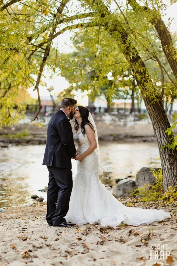 A couple is captured in a romantic moment at the riverside wedding venue of Hôtel Château Joliette, a renowned destination featured in the wedding venues category of Mariage Elegant, Canada. The bride is elegantly dressed in a white lace gown with a long veil, and the groom is wearing a classic black suit. They are standing on a sandy riverbank, surrounded by lush green trees with leaves gently swaying in the breeze. The tranquil river adds a picturesque backdrop to this intimate scene, ideal for an outdoor wedding ceremony.
