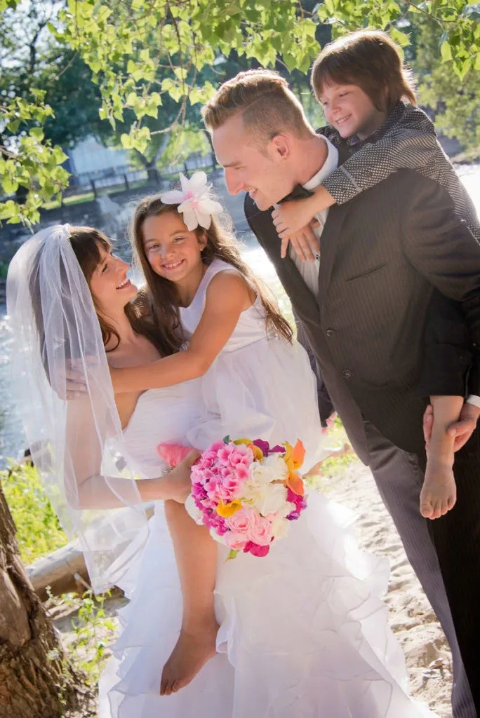 A joyful wedding scene at Hôtel Château Joliette, a prominent wedding venue in Canada, features a newlywed couple with their two young children. The bride, in a flowing white gown and veil, holds a vibrant bouquet of pink and white flowers. She is smiling warmly at her daughter, who is dressed in a white dress with a flower in her hair. The groom, in a dark suit, carries their son on his shoulders, sharing a happy moment by a riverside under the shade of green leaves. This image encapsulates a family-friendly celebration at a picturesque wedding venue.