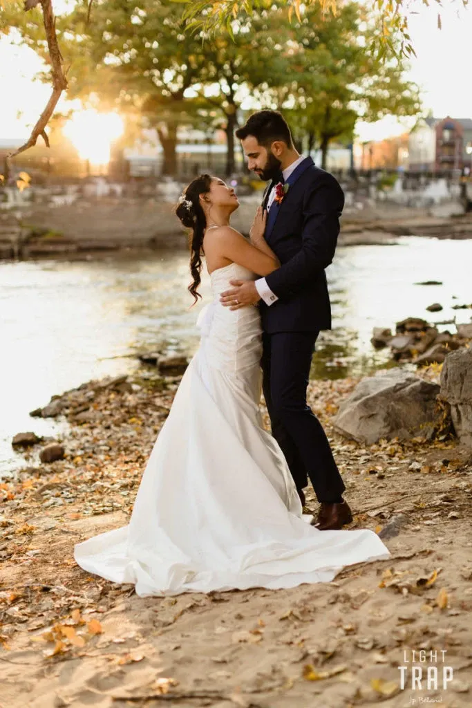A bride and groom stand on a serene riverbank during sunset, surrounded by autumn leaves. The bride is wearing an elegant white wedding gown, and the groom is dressed in a dark suit. They are gazing into each other's eyes, creating a romantic and intimate moment. This picturesque setting highlights Hôtel Château Joliette as an ideal wedding venue for couples seeking a natural and beautifully scenic backdrop for their special day. The soft lighting and tranquil water add to the enchanting atmosphere perfect for a wedding celebration.