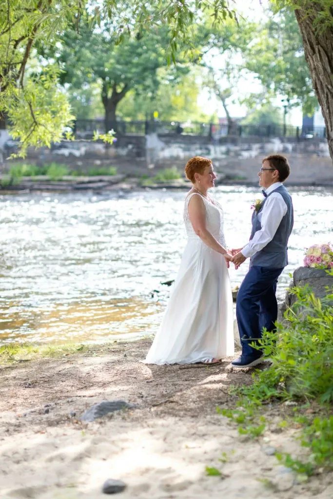 A happy couple stands hand in hand by a picturesque riverside setting at Hôtel Château Joliette, a wedding venue in Canada. The bride is wearing a flowing white gown, and the groom is in a stylish suit. Sunlight filters through the trees, casting a gentle glow on the tranquil water, creating a romantic and serene atmosphere for a wedding at this elegant venue.