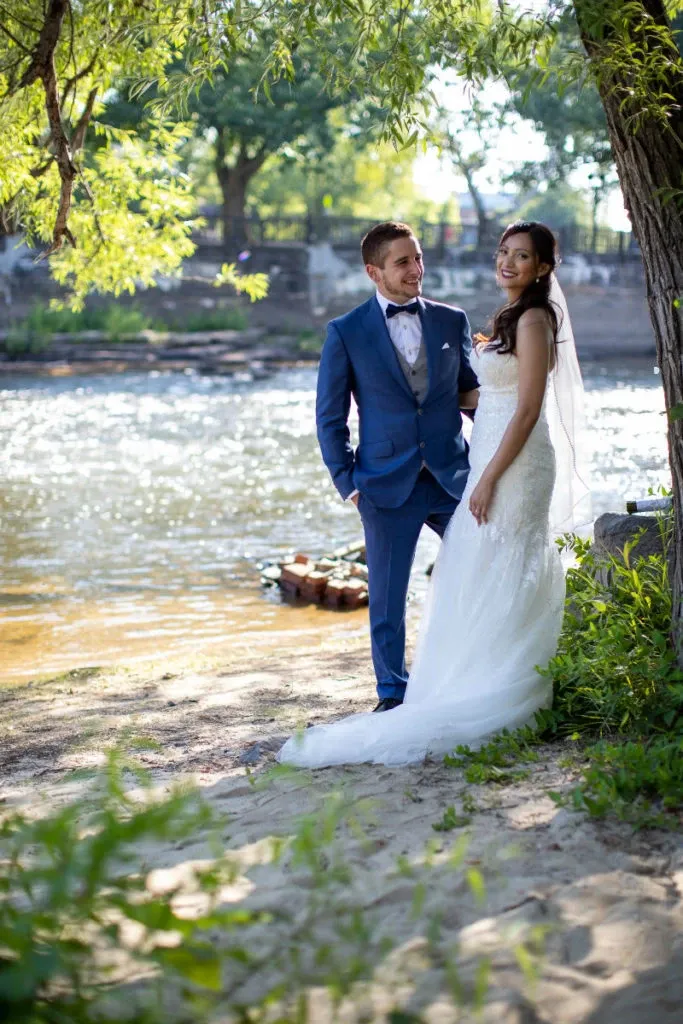 A bride and groom stand on a sandy riverbank under a canopy of green trees at Hôtel Château Joliette, a wedding venue in Canada. The bride is wearing a white lace wedding gown with a long veil, and the groom is dressed in a formal blue suit with a bow tie. They are looking at each other and smiling, capturing a moment of joy and romance. The serene river flows gently in the background, creating a picturesque and intimate setting for a wedding celebration.