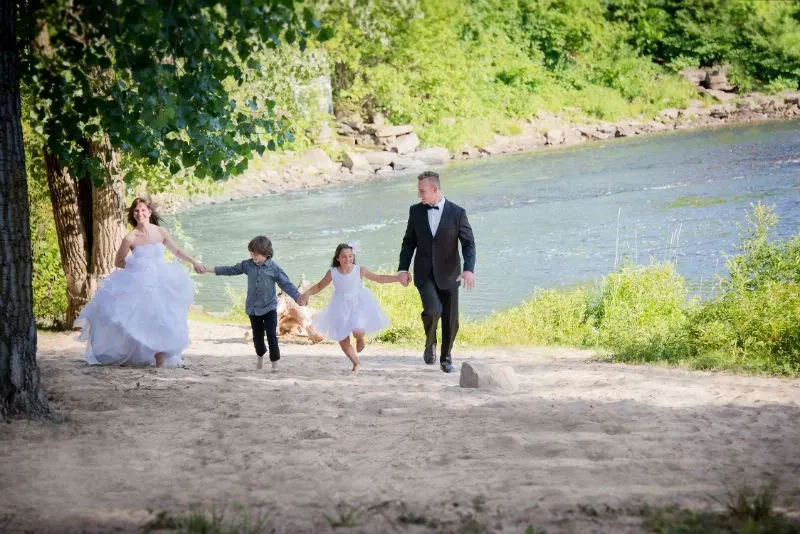 The image shows a joyful scene of a bride and groom holding hands with two children, walking along a sandy riverbank. The bride is wearing a flowing white wedding gown, and the groom is dressed in a formal black suit with a bow tie. The children, a boy and a girl, are dressed in festive attire suitable for the occasion. The lush greenery and tranquil river in the background suggest a picturesque outdoor setting. This image is representative of the wedding venues category and features Hôtel Château Joliette, highlighting its beautiful natural surroundings perfect for wedding celebrations.