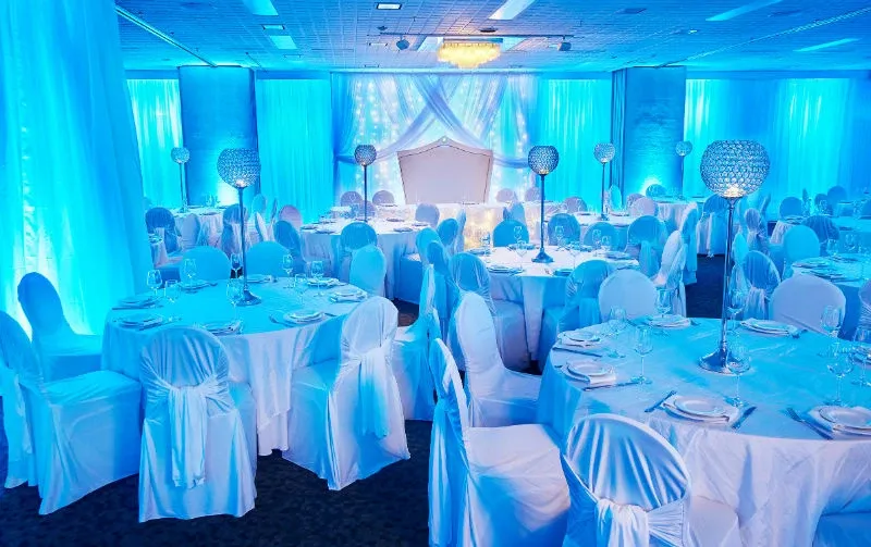 The image showcases a beautifully decorated wedding venue at Hôtel Château Joliette, highlighting its suitability for elegant wedding celebrations. The reception hall features round tables draped with white tablecloths and chairs adorned with white covers and sashes. The ambient blue lighting creates a romantic atmosphere, accentuated by decorative floor-to-ceiling drapes. Crystal centerpieces are placed on each table, adding a touch of glamour. The backdrop features a softly lit, draped archway, perfect for wedding ceremonies or speeches, completing the sophisticated setting for this wedding venue.