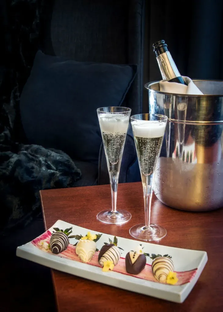The image features a romantic setup at Hôtel Château Joliette, a wedding venue in Canada. Two elegant champagne flutes filled with sparkling wine are placed on a wooden table next to a chilled bottle in a silver ice bucket, creating a celebratory atmosphere. In the foreground, a decorative white plate displays a selection of gourmet chocolate-covered strawberries, garnished with small edible flowers. The setting exudes sophistication, making it ideal for an intimate wedding celebration or reception at the venue.