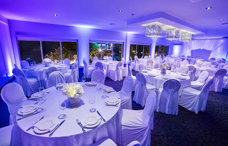 The image showcases a beautifully decorated wedding venue at Hôtel Château Joliette, categorized under Wedding Venues on Mariage Elegant, Canada. The room is elegantly lit with ambient blue lighting, creating a romantic atmosphere. Round tables are covered with pristine white tablecloths, each adorned with a centerpiece of white flowers. Chairs are draped in white covers, enhancing the sophisticated look. The ceiling features modern, sparkling chandeliers that add a touch of glamour to the setting. Large windows line one side of the room, offering a view of greenery outside. The overall scene is perfect for an elegant and memorable wedding celebration.