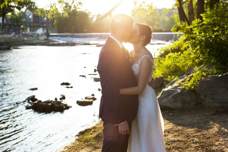 A bride and groom share a romantic kiss beside a serene river, surrounded by lush greenery, with the sun setting gently behind them. The bride is elegantly dressed in a flowing white gown, while the groom wears a classic dark suit. This picturesque scene is captured at the Hôtel Château Joliette, a stunning wedding venue in Canada, known for its natural beauty and intimate setting, perfect for celebrating love and beginning a new life together.