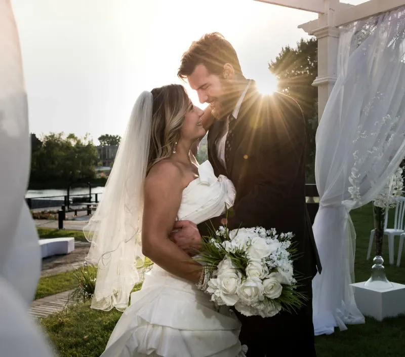 A bride and groom share an intimate moment under a gazebo at Hôtel Château Joliette, representing their wedding venue category. The bride, in an elegant white gown with a long veil, holds a bouquet of white roses. The groom is dressed in a classic black suit. Sunlight casts a warm glow on their faces, enhancing the romantic atmosphere. The gazebo is adorned with sheer white drapes, adding to the picturesque setting of a riverside garden, ideal for a wedding ceremony.