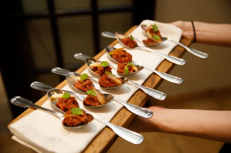 A server presents a wooden board with several elegantly arranged spoons containing bite-sized appetizers, garnished with fresh herbs. This sophisticated setup highlights the culinary offerings at Hôtel Château Joliette, showcasing its expertise as a premier wedding venue in Canada. The attention to detail in presentation reflects the venue’s commitment to providing a memorable dining experience for wedding guests.