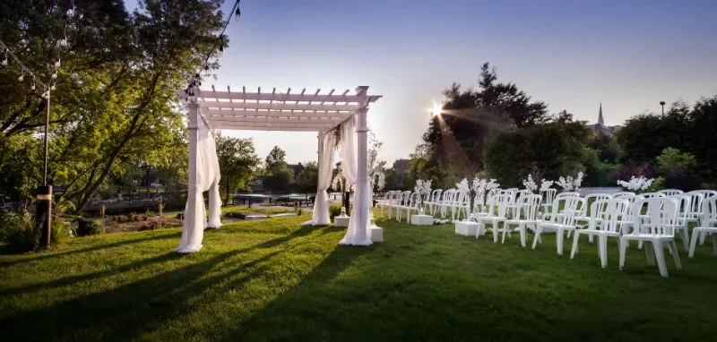 The image showcases the enchanting outdoor wedding venue at Hôtel Château Joliette, part of the Wedding Venues category on Mariage Elegant, Canada. Set in a lush green garden, the space features a charming white pergola adorned with flowing white drapes. Rows of elegant white chairs are arranged neatly on the lawn, ready for guests. The scene is beautifully lit by soft, ambient lighting, with strings of lights hanging above, creating a romantic atmosphere as the sun sets in the background. Perfect for couples seeking a picturesque outdoor ceremony.