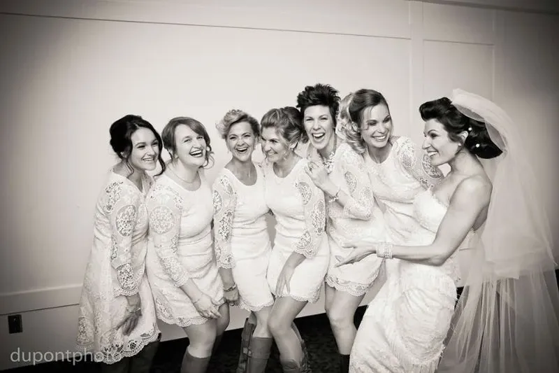 A group of seven women, including a bride, is joyfully gathered together in a hallway. The bride is wearing an elegant lace wedding gown with a long veil, while the six bridesmaids are dressed in matching short lace dresses. Their laughter and camaraderie create a lively and cheerful atmosphere. This scene, captured at Hôtel Château Joliette, exemplifies the venue’s suitability for intimate and joyous wedding celebrations, highlighting its charm as an ideal wedding venue in Canada.