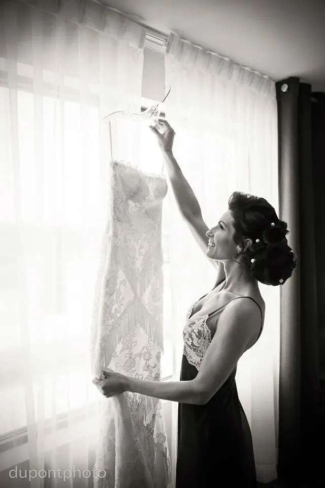 A bride joyfully admires a lace wedding dress hanging by a window in a well-lit room at Hôtel Château Joliette. She is preparing for her big day, capturing a moment of excitement and anticipation. This image highlights the elegance and intimate setting offered by Hôtel Château Joliette, a prime choice for wedding venues in Canada.