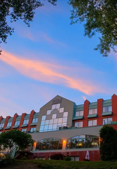 The image showcases Hôtel Château Joliette, a premier wedding venue featured on Mariage Elegant in Canada. The hotel's façade is visible with a modern, multi-story design featuring large windows reflecting the evening sky. The exterior is surrounded by lush greenery, enhancing its picturesque setting. The sky above is painted in shades of blue and pink, suggesting a serene, romantic atmosphere ideal for weddings.